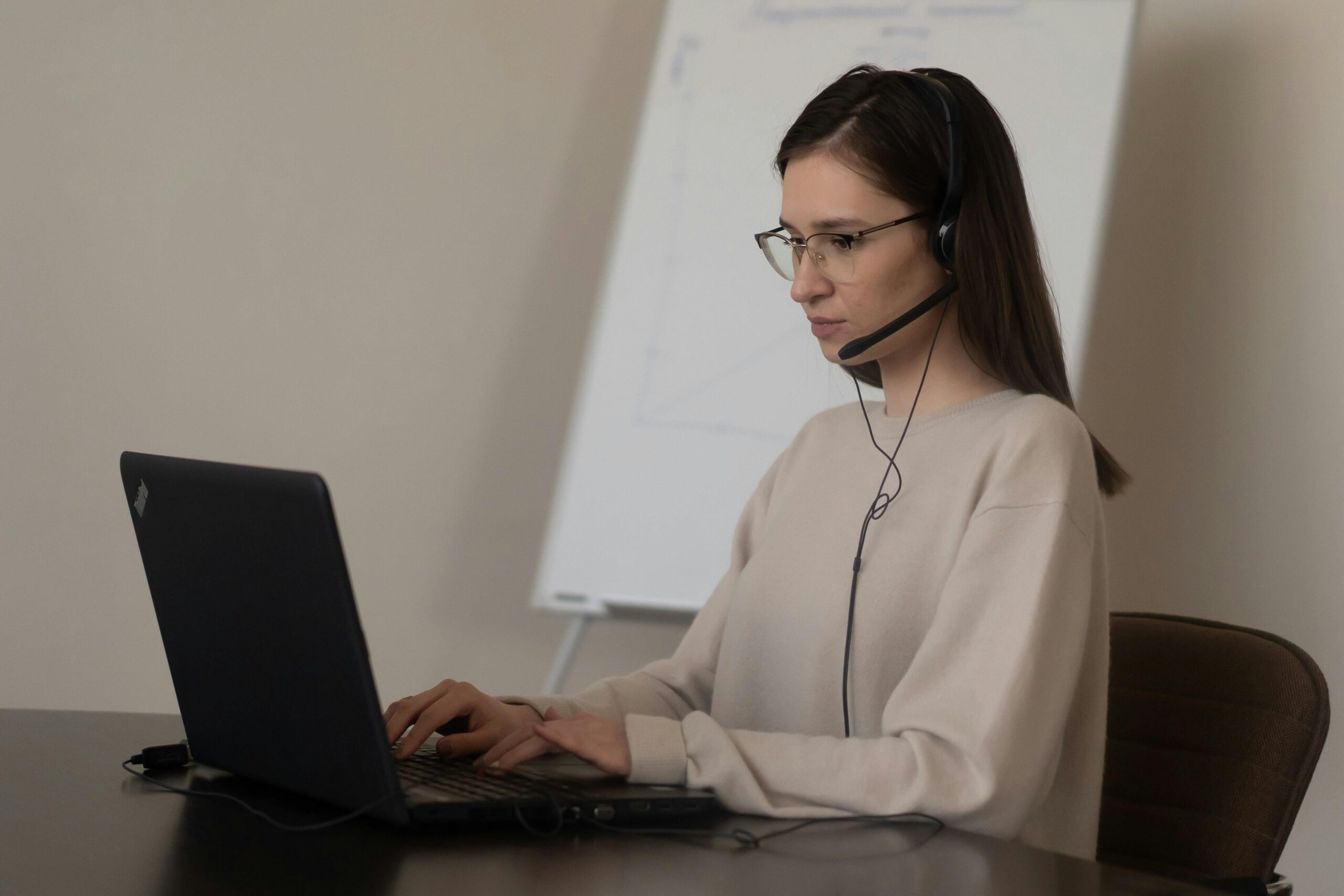 Serious young woman in casual outfit with eyeglasses sitting on chair at table and working on laptop with headphones with microphone near whiteboard in bright workplace